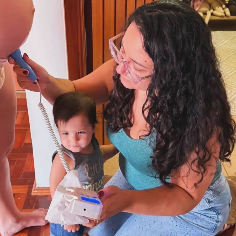 Arielle Matos ausculta os batimentos do bebê com sonar, ao lado de uma criança curiosa, durante atendimento em casa
